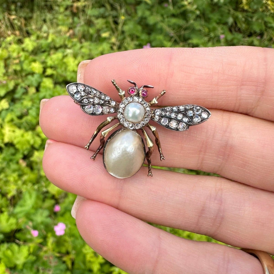 Victorian bee brooch featuring cut diamonds, pearls, and rubies, held in hand against a green background.